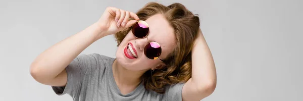 Γοητευτική νεαρή κοπέλα σε ένα γκρι μπλουζάκι σε γκρι φόντο. Χαρούμενο κορίτσι σε στρογγυλά γυαλιά — Φωτογραφία Αρχείου
