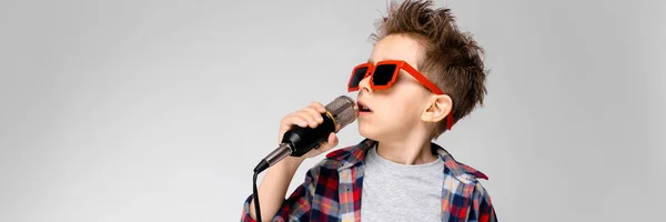 Ένα όμορφο αγόρι σε ένα καρό πουκάμισο, γκρι πουκάμισο και τζιν στέκεται σε γκρι φόντο. Ένα αγόρι που φορώντας γυαλιά ηλίου. Κόκκινα μαλλιά αγόρι που τραγουδάει στο μικρόφωνο — Φωτογραφία Αρχείου