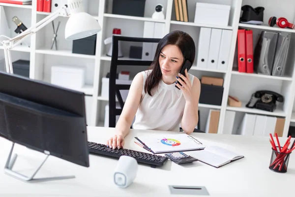 Молодая девушка сидит в офисе за компьютерным столом, разговаривает по телефону и работает с документами . — стоковое фото