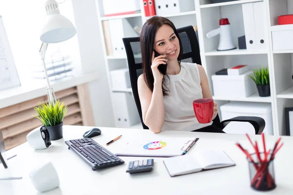 Una joven sentada en la oficina en el escritorio de la computadora, hablando por teléfono y sosteniendo una taza roja . — Foto de Stock