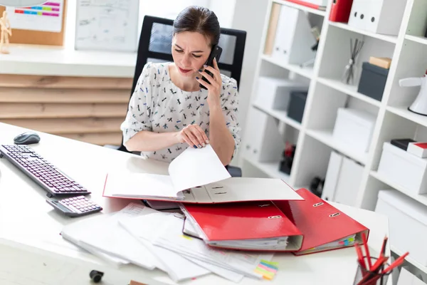 Uma jovem sentada no escritório na mesa do computador, trabalhando com documentos e falando ao telefone . — Fotografia de Stock