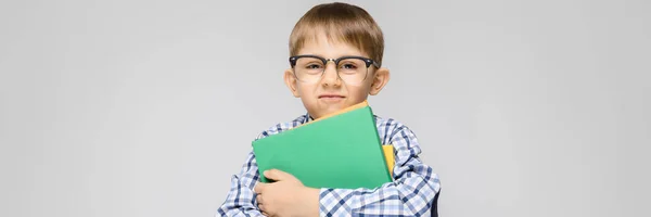 Um menino encantador com uma camisa de vkletchatoy e jeans leves fica em um fundo cinza. O menino segura em suas mãos uma pasta multicolorida com documentos — Fotografia de Stock