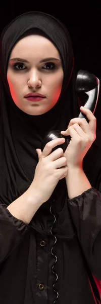 Porträt einer schönen, ernsthaft verängstigten jungen Muslimin, die einen schwarzen Hijab trägt und ein Telefon hält, um als Wahlkonzept auf schwarzem Hintergrund anzurufen — Stockfoto