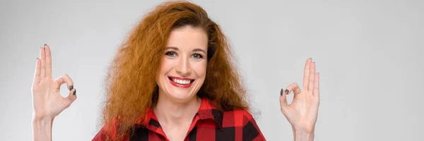 Mosolyogva mutatja az ok jele, a szürke háttér gyönyörű vörös hajú boldog fiatal nő portréja — Stock Fotó