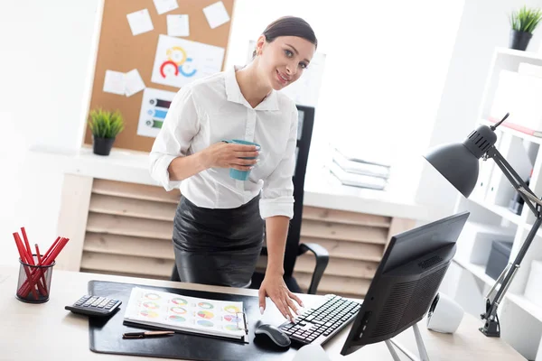 Una joven de pie en la oficina, sosteniendo una taza y escribiendo en el teclado . — Foto de Stock