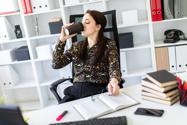 Una joven sentada en la oficina en la mesa de la computadora y sosteniendo un vaso de café . — Foto de Stock