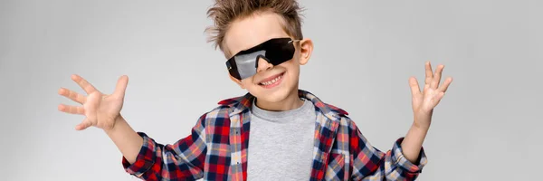 Ένα όμορφο αγόρι σε ένα καρό πουκάμισο, γκρι πουκάμισο και τζιν στέκεται σε γκρι φόντο. Το αγόρι με μαύρα γυαλιά ηλίου. Το αγόρι εξάπλωση τα χέρια του προς τις πλευρές. — Φωτογραφία Αρχείου