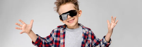 Un chico guapo con una camisa a cuadros, camisa gris y jeans está parado sobre un fondo gris. El chico con gafas de sol negras. El chico extendió sus manos a los lados. . — Foto de Stock