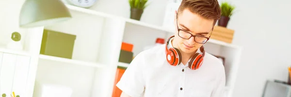 A young man in glasses stands near a computer desk. A young man draws a marker on a magnetic board. On the neck, the guys headphones hang.