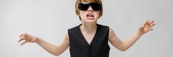 Retrato de adorable niño divertido de pie en el estudio con gafas de sol que muestran la agresividad ira con ganas de rascarse en el fondo gris —  Fotos de Stock