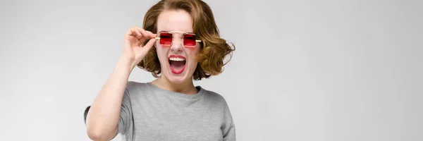 Encantadora joven con una camiseta gris sobre un fondo gris. Chica en gafas de ojos cuadrados rojos . —  Fotos de Stock