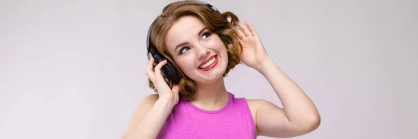 Okouzlující mladá dívka v růžových šatech na šedém pozadí. Mladá dívka se sluchátky s úsměvem — Stock fotografie