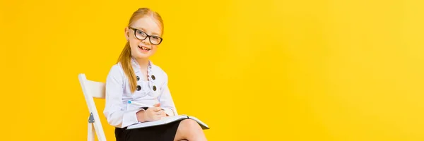 Ragazza con i capelli rossi su sfondo giallo. Una ragazza affascinante in occhiali trasparenti si siede su una sedia bianca e prende appunti in un quaderno . — Foto Stock