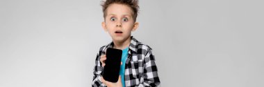 Yakışıklı bir çocuk bir ekose gömlek, mavi tişört ve kot gri bir arka plan üzerinde duruyor. Çocuk bir telefon tutarak