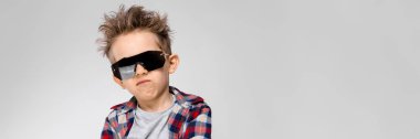 Yakışıklı bir çocuk bir ekose gömlek, gri gömlek ve pantolon gri bir arka plan üzerinde duruyor. Siyah güneş gözlüklü çocuk. Çocuğun kollarını göğsüne üzerinde katlanmış.