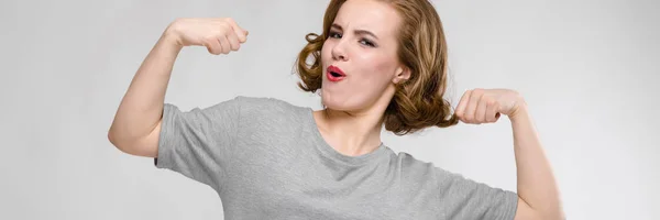 Menina encantadora em uma camiseta cinza em um fundo cinza. Menina mostrando músculos — Fotografia de Stock