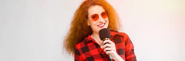 Portrait de belle rousse heureuse jeune femme en lunettes de soleil souriant tenant microphone sur fond gris — Photo