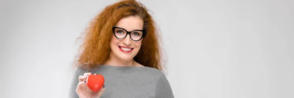 Jovem encantadora com cabelo vermelho. Uma rapariga com uma camisola cinzenta. Menina em um fundo cinza. Uma jovem com óculos está segurando um coração vermelho — Fotografia de Stock