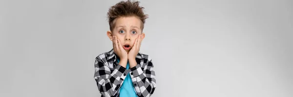 Un beau garçon en chemise à carreaux, chemise bleue et jean se tient sur un fond gris. Le garçon tient ses mains sur ses joues — Photo