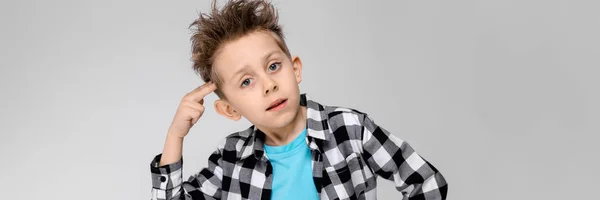 A handsome boy in a plaid shirt, blue shirt and jeans stands on a gray background. The boy put two fingers to his temple — Stock Photo, Image