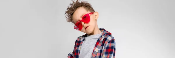 Um menino bonito em uma camisa xadrez, camisa cinza e jeans fica em um fundo cinza. Um rapaz de óculos de sol vermelhos. O rapaz pôs a mão no braço . — Fotografia de Stock