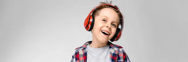 Een knappe jongen in een plaid shirt, grijs shirt en spijkerbroek staat op een grijze achtergrond. Een jongen in rode hoofdtelefoon. De jongen heeft zijn handen op zijn buik. De jongen lacht. — Stockfoto