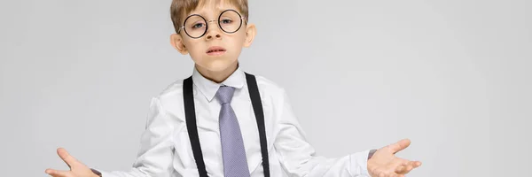 Um menino encantador com uma camisa branca, suspensórios, uma gravata e jeans leves fica em um fundo cinza. O menino estendeu as mãos em ambas as direções — Fotografia de Stock