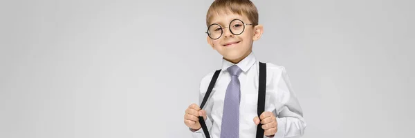 Um menino encantador com uma camisa branca, suspensórios, uma gravata e jeans leves fica em um fundo cinza. o rapaz de óculos puxou os suspensórios — Fotografia de Stock