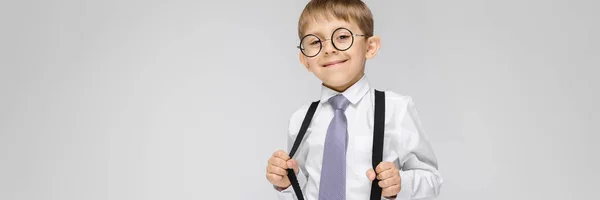 Un niño encantador con una camisa blanca, tirantes, corbata y pantalones vaqueros ligeros se levanta sobre un fondo gris. el chico con gafas tiró de los tirantes —  Fotos de Stock