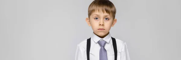 Um menino encantador com uma camisa branca, suspensórios, uma gravata e jeans leves fica em um fundo cinza — Fotografia de Stock