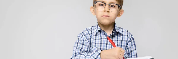 Um menino encantador com uma camisa de vkletchatoy e jeans leves fica em um fundo cinza. O rapaz tem um caderno e uma caneta nas mãos. . — Fotografia de Stock