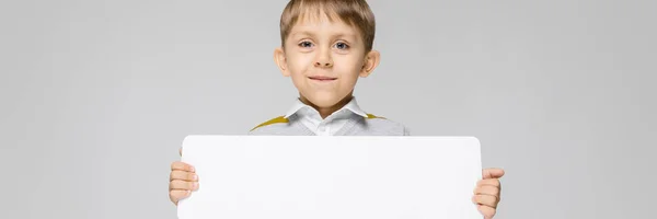 Un niño encantador con una camisa blanca, camiseta de tirantes a rayas y pantalones vaqueros ligeros está sobre un fondo gris. El niño sostiene un afiche rectangular blanco —  Fotos de Stock