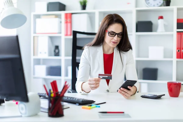 Una joven sentada en la oficina a la mesa con una tarjeta bancaria y un teléfono . — Foto de Stock