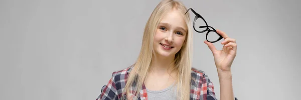 Charmantes Teenager-Mädchen mit langen blonden Haaren hält eine Brille in der Hand. ein Teenager-Mädchen in kariertem Hemd und grauem T-Shirt. — Stockfoto