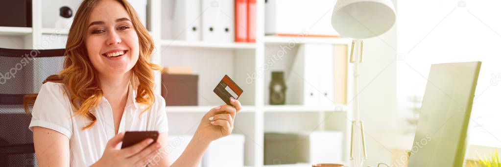 Beautiful young girl sits in the office, holds a bank card and phone in her hand.