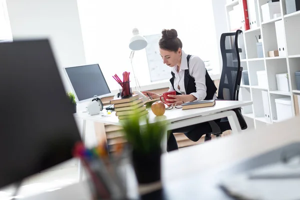 En ung flicka sitter vid ett bord på kontoret, har en röd kopp och penna i händerna och fungerar med dokument. — Stockfoto