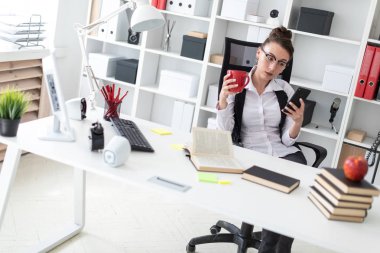 Genç bir kız ofiste bilgisayar masasında oturuyor ve bir kırmızı Kupası ve telefonu tutuyor.