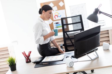 Genç bir kız ofiste bir tabloda bir fincan tutarak ve bir bilgisayar ve belgeleri çalışma oturdu.