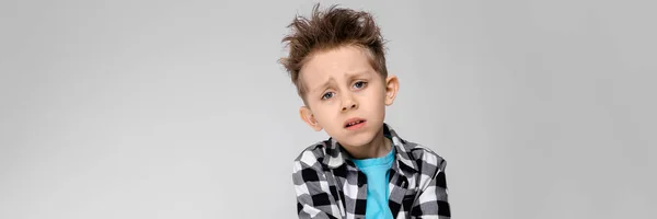 A handsome boy in a plaid shirt, blue shirt and jeans stands on a gray background. The boy folded his arms over his chest. The boy is unhappy. The boy shows his hand — Stock Photo, Image