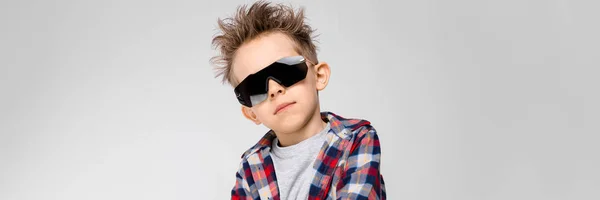 En stilig pojke i en rutig skjorta, grå skjorta och jeans står på en grå bakgrund. Pojken i den svarta solglasögonen. Pojken viks armarna över bröstet — Stockfoto