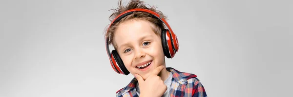 Ένα όμορφο αγόρι σε ένα καρό πουκάμισο, γκρι πουκάμισο και τζιν στέκεται σε γκρι φόντο. Ένα αγόρι στο κόκκινο ακουστικά. Το αγόρι κρατά ένα χέρι στο πηγούνι του. — Φωτογραφία Αρχείου