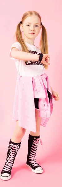 Chica con colas rojas sobre un fondo rosa. Una chica encantadora con ropa deportiva brillante mira su reloj . —  Fotos de Stock