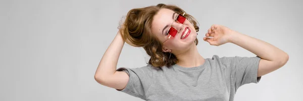 Γοητευτική νεαρή κοπέλα σε ένα γκρι μπλουζάκι σε γκρι φόντο. Κόκκινη Πλατεία-eyed γυαλιά κοριτσιών. — Φωτογραφία Αρχείου