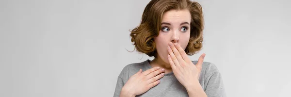 Charmantes junges Mädchen in einem grauen T-Shirt vor grauem Hintergrund. das Mädchen bedeckte ihren Mund mit der Hand — Stockfoto