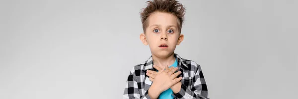 Um menino bonito em uma camisa xadrez, camisa azul e jeans fica em um fundo cinza. O rapaz dobrou as palmas das mãos no peito — Fotografia de Stock