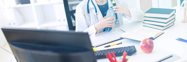 A beautiful young girl in a white robe sits at a computer desk, holds a marker in her hand and opens a bottle of water.