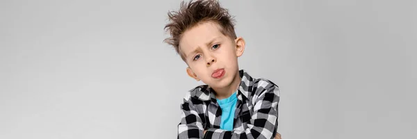 A handsome boy in a plaid shirt, blue shirt and jeans stands on a gray background. The boy folded his arms over his chest. The boy shows the language — Stock Photo, Image