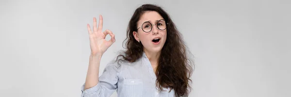 Retrato de uma jovem de óculos. Menina bonita em um fundo cinza. A menina mostra o sinal dos okkes . — Fotografia de Stock