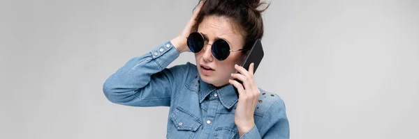 Jeune fille brune en lunettes rondes. Les cheveux sont rassemblés dans un chignon. Une fille avec un téléphone noir. La fille parle au téléphone. . — Photo