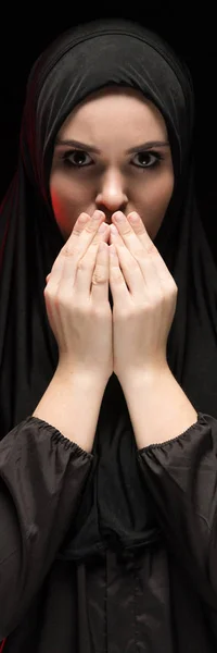 Retrato de una hermosa joven musulmana seria usando hiyab negro con las manos cerca de su cara como concepto de oración sobre fondo negro — Foto de Stock